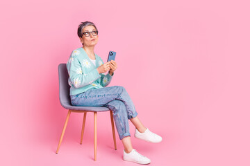 Stylish mature woman posing with smartphone seated against a vivid pink background showcasing modern casual spring fashion.