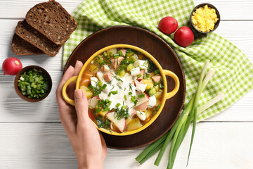 Female hand with bowl of tasty okroshka, ingredients and bread on white wooden background, closeup