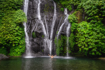 A men enjoys stunning Banyumala Twin Waterfalls that cascades into clear pool surrounded by vibrant...