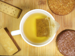 Closeup overhead shot of biscuits surrounding a white cup of hot water and an infusing tea bag containing natural, organic herbs and seeds.