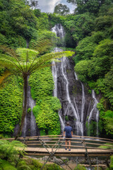 Men on a bridge enjoys stunning Banyumala Twin Waterfalls that majestically cascades down the rocky cliffs, surrounded by vibrant, lush greenery, offering a tranquil tropical escape, Bali, Indonesia