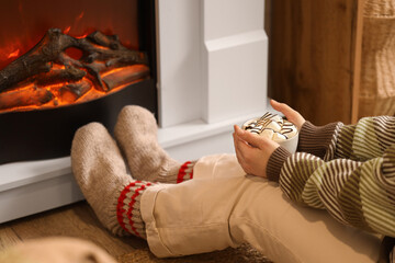 Young woman with cup of cocoa sitting near fireplace at home, closeup