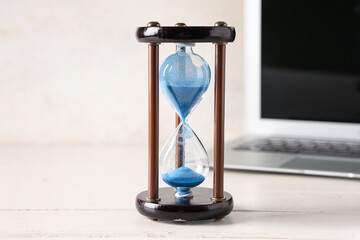 Stylish hourglass with blue sand and laptop on table at workplace