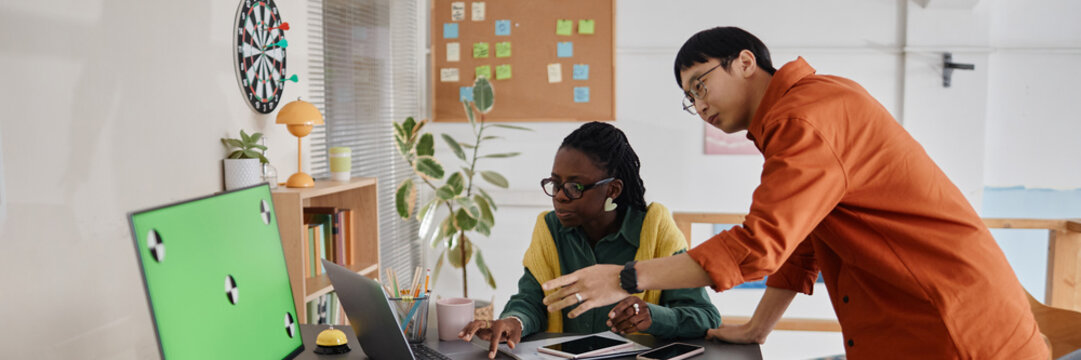 Two coworkers collaborating on project at desktop computer while discussing ideas surrounded by office decor and plants. Green screen visible on monitor in foreground - Powered by Adobe