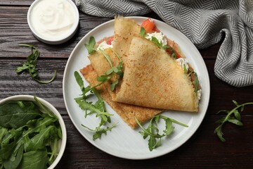 Delicious crepes with salmon, cream cheese and arugula on wooden table, flat lay