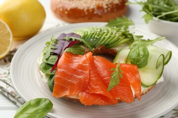 Delicious bagel with salmon, cream cheese, cucumber and avocado on white wooden table, closeup