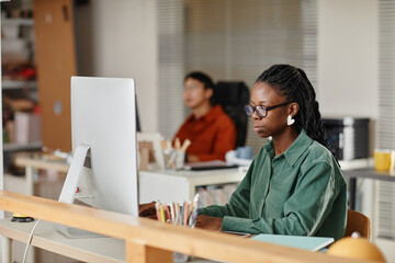 Young professional focused on computer screen in modern office environment showing serious expression. Colleagues and office supplies are visible in background
