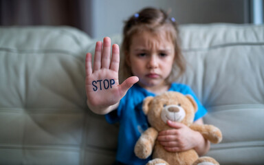 An afraid, sad little girl with stop gesture, "stop" written on her palm at home. Rejection of domestic violence home, child abuse, discrimination, bullying concept.