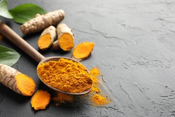 Aromatic turmeric powder in spoon, fresh roots and leaves on black table, closeup. Space for text