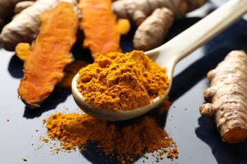 Aromatic turmeric powder in spoon and fresh roots on blue plate, closeup