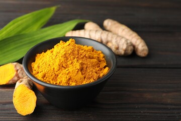 Aromatic turmeric powder in bowl, fresh roots and leaves on wooden table, closeup. Space for text
