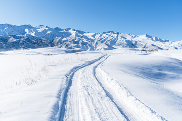 Snow track on a winter field going to the mountain ranges covered with snow and blue clear sky. Sunny winter day in the mountains.