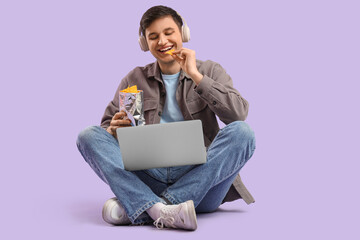 Young man with laptop eating tasty nachos and listening to music on lilac background