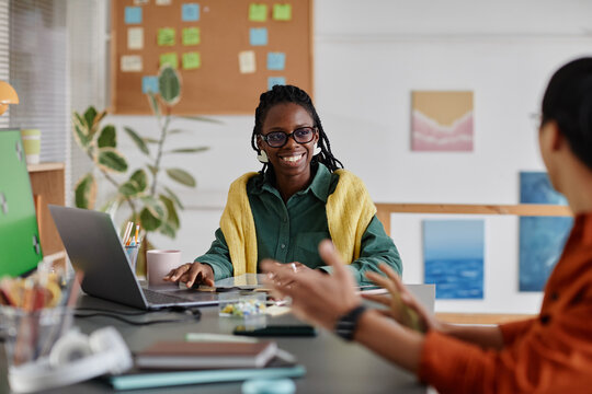Smiling woman sitting in office environment with colleague, engaging in casual conversation. Books, laptop, and plant seen in background, providing a warm, professional setting