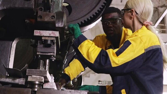 Two male and female factory mechanics operate metal stamping machines together in a factory in the automotive parts manufacturing industry, turning and metal stamping.