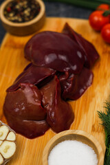 Close-up of raw chicken liver on a wooden board surrounded in out-of-focus garlic, tomatoes, seasonings and salt in wooden bowls and herbs on a dark background. The concept of healthy eating, cooking