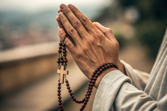 Close-up of praying hands holding a wooden rosary with a cross outdoors