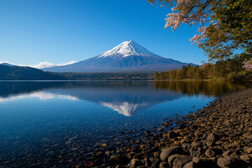 Fototapeta premium stunning Mount Fuji reflects beautifully in calm waters, surrounded by cherry blossoms and clear blue sky, creating serene and picturesque landscape