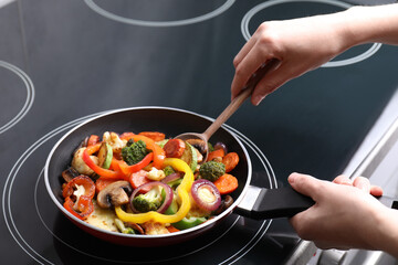 Woman cooking vegetables and mushrooms in frying pan on cooktop indoors, closeup