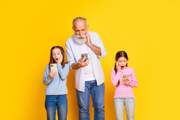 Elderly man and two young girls sharing a surprising moment while using smartphones, enjoying quality time together against a bright yellow studio background