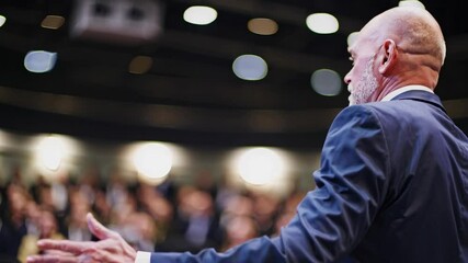 Senior businessman in a suit delivers a powerful speech at a large conference, engaging with the audience through expressive hand gestures