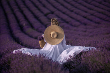 Lavender Fields Woman France: A woman in a white dress and straw hat sits amidst blooming purple lavender fields in Provence, France, during summer, for aesthetic photography.