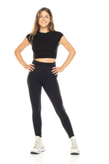 Fototapeta premium Young smiling woman in black crop top and high-waisted leggings. Long brown hair. Full-body shot against white background. Wearing white sneakers. Neutral expression