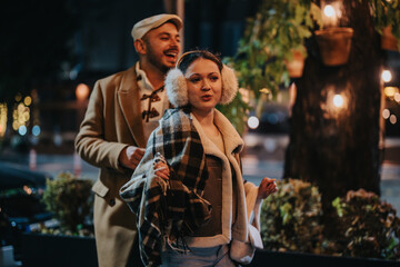 A cheerful couple walks through a warmly lit outdoor area, wrapped in cozy winter clothing. They seem to be enjoying a pleasant evening, surrounded by festively decorated plants and soft lighting.