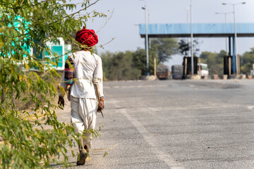 shepherd in traditional dress with turban and stick and walking at tarmac road near village at day