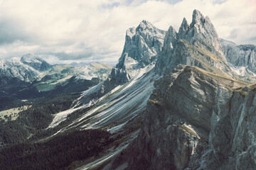 A vintage style image of the Seceda mountain range in the Dolomites, Italy, with rugged cliffs and forested valleys. A muted film effect enhances the atmospheric tone