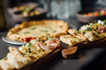 Italian antipasti platter featuring aged cheeses (parmesan, pecorino, chevre), prosciutto, olives, and focaccia, in front of pizza. Restaurant scene with shallow depth of field, soft bokeh