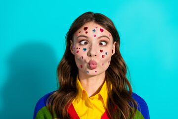 Young woman making a funny face with colorful stickers on her face, wearing a vibrant rainbow sweater against a teal background