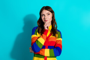 Stylish young woman with thoughtful expression in rainbow sweater on turquoise background,...