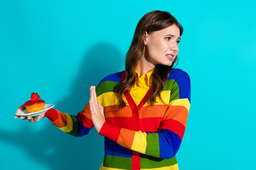 Stylish young woman in a rainbow cardigan refusing a dessert against a teal background