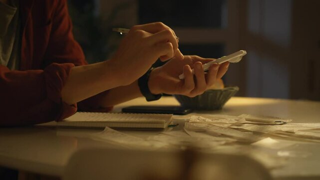 A young adult sits at a table, taking notes while checking their phone amidst scattered receipts. The dimly lit space creates a focused atmosphere for managing finances late at night