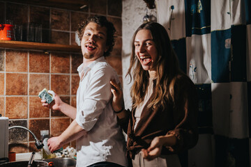 Two young adults happily spend time together in a kitchen environment, washing dishes and sharing laughter, showing companionship and enjoyment of casual moments.