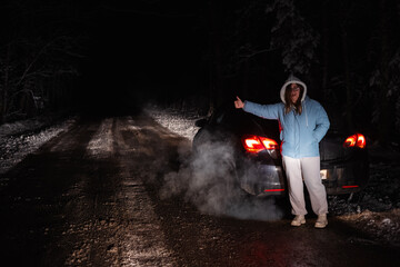 Obraz premium Woman stands next to her broken-down car on a snowy forest road at night, signaling for help. The cold winter scene is illuminated by the car's rear lights and rising steam.