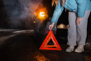 On a dark winter road, a woman places a reflective warning triangle beside her broken-down car. The...