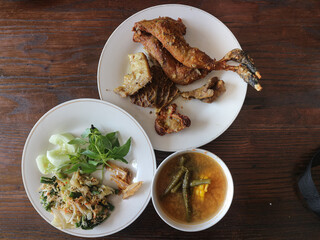 A plate of fried chicken and offal, a plate of urap vegetables or sayur urap, and a bowl of sour vegetables or sayur asem, served on a brown table