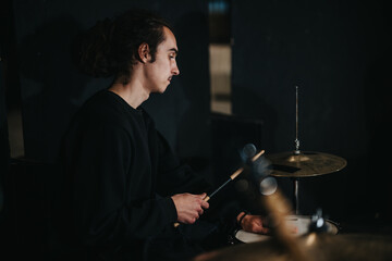 A young man with long hair plays drums with passion in a cozy studio setting, highlighting the...