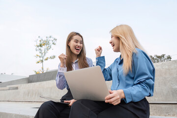 Two businesswomen outdoors joyfully celebrating success with laptops.