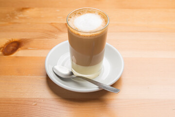 Coffee latte macchiato in a tall clear glass with a spoon on a saucer on a wooden table