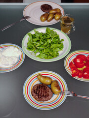 Grilled raw burger patties beside boiled potatoes, green salad, tomato slices, herbal tea on glass table