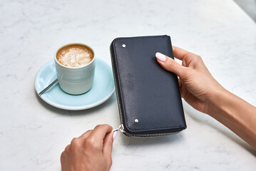 Hand holding black leather wallet, opening zipper on wallet at cafe table, coffee beside, stylish accessory in hand concept