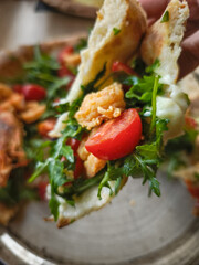 Hand holding Close up of crispy italian flatbread layering fresh arugula, ripe cherry tomatoes, melting cheese on rustic wooden surface