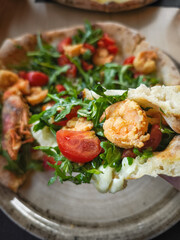 Hand holding Gourmet pizza featuring succulent shrimp, peppery arugula, and ripe cherry tomatoes on rustic surface in aosta, italy