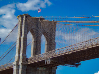 Fototapeta premium A striking view of the Brooklyn Bridge in New York City, showcasing its iconic stone towers and intricate web of steel cables against a vibrant blue sky. The American flag waves proudly atop the struc