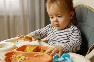 A curious baby sits in a high chair, engaged with a colorful tray of food. The little one plays...