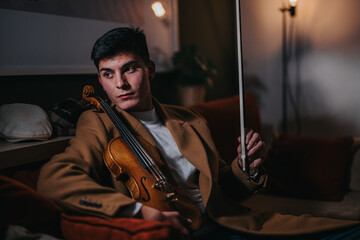 A young man relaxing while holding a violin and bow, sitting in a cozy room. The ambient lighting...