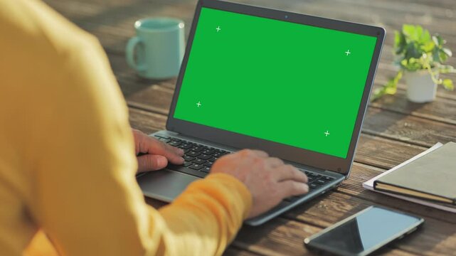 man typing at the laptop with green screen back view,person types at the computer outdoor on the wooden table rear view,chroma key display